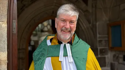 Barker stood in front of the entrance to a church. He has a grey beard and short grey hair. He is wearing traditional religious clothing which is white, yellow and green. 