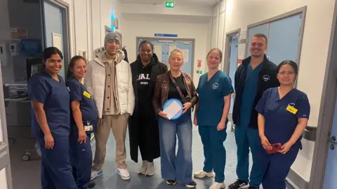 A line of nurses in blue uniform, with Mr Sarpong, his girlfriend and his mum standing in the middle of them. They are in what looks like a hospital corridor.