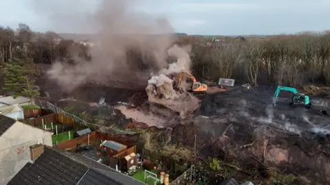 Drone shot over the roof of some houses showing digger on burning embankment.