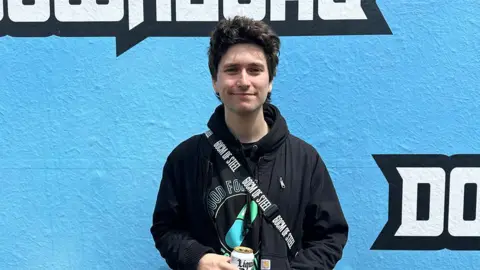 Lucas Woodland from pop-punk band Holding Absence stood in front of a blue wall with the word Download written across it. He is wearing all black, smiling and holding a can of water.