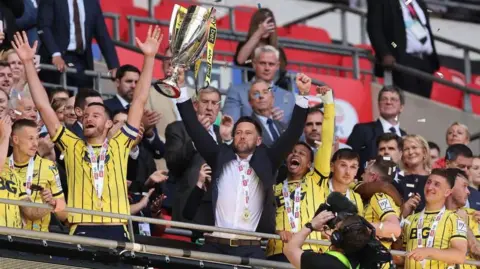 Getty Images Oxford United manager Des Buckingham lifts the League One play-off final trophy while his team celebrates around him.