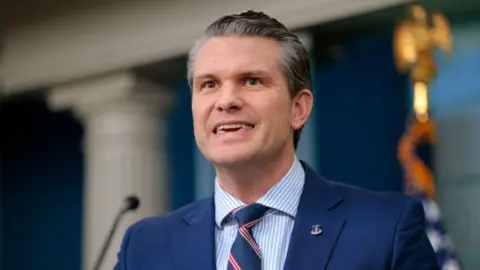 US Defence Secretary Pete Hegseth speaks at a news conference. He wears a blue suit, striped blue and white shirt and a navy and red tie.