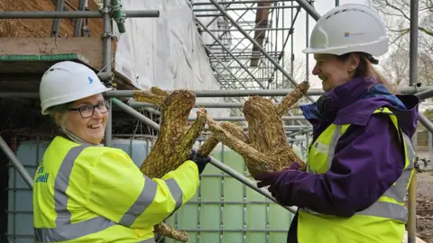 Historic England Archive Two women wearing high-viz and hard hats hold thatched hares.