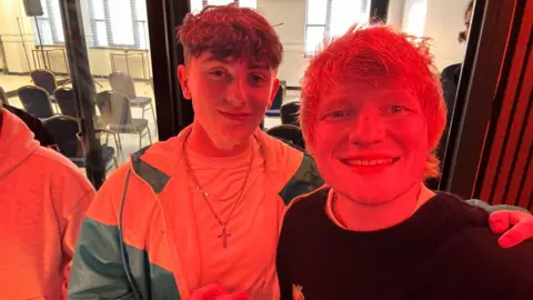 Dylan Magee-Brownlee Ed Sheeran is posing for a selfie with a young man. They are in a studio with a red light shining. 