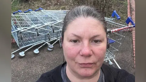 Kirsty Knight Kirsty Knight standing in front a fallen-down line of trolleys - taking a selfie- wearing a grey T-shirt and a black cardigan