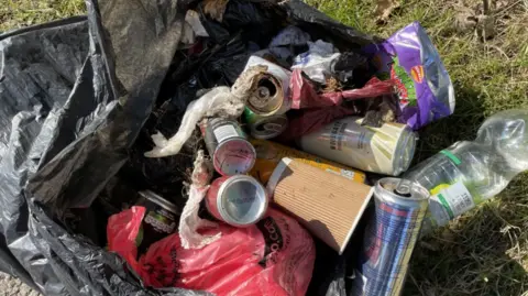 An open black bin bag containing various items of rubbish, including cans, coffee cups and crisp packets. It is laid out on an area of grass.