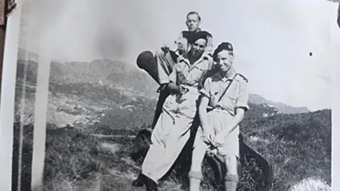 Joseph Douglas Cheshire Joseph Douglas Cheshire sitting on a cannon with two fellow soldiers on "Hong Kong Peak" with mountains behind them. All wearing military uniforms and two of them wearing berets