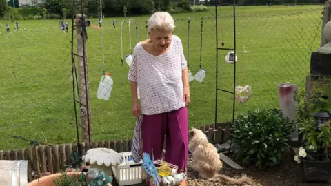 Louise Fenner Madeline is standing at the end of her garden with her cream coloured cat. She has bottles of water hanging over her wired fence. Madeline has grey hair and is wearing a pink oversized t-shirt with purple trousers. 
