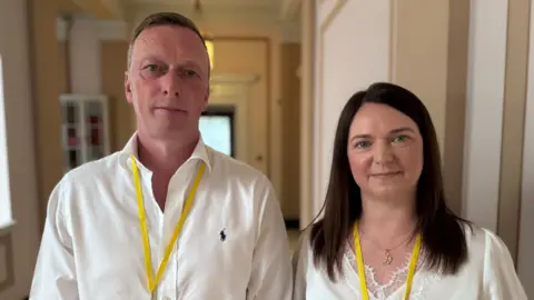 Neville and Joanne Reid. Neville has short brown hair, wearing a white shirt and yellow lanyard. Joanne has dark shoulder length hair, wearing a sliver moon necklace, white top and yellow lanyard.