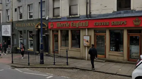 A high street with a red sign outside in gold writing which reads 'Merrie England coffee shops'.