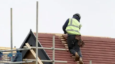 A construction worker in a hi-vis jacket is on a roof that is being built. Some scaffolding is visible on the left.