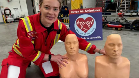 Rhiannon Roderick crouches next to two training manikins. One is a male torso and the other is a female torso. She is wearing a red paramedic jumpsuit. In the background there is a Devon Air Ambulance Sign. 
