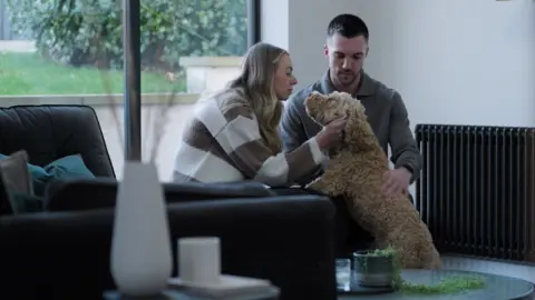 Stewarts Solicitors A woman with blonde hair and brown and white stripy jumper, pets a brown dog which has its paws on her lap. A man with dark hair and brown jumper is next to them