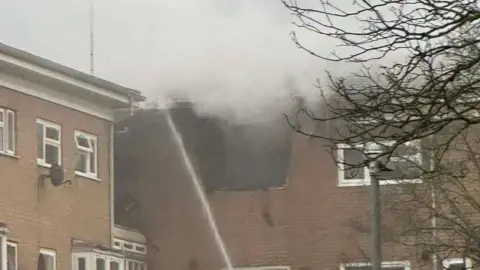 Lucas A close up of a flat with a gaping hole in the roof and wall, and smoke emitting from the hole. A stream of water is pointed at the hole.