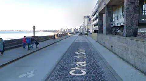 A seafront promenade with a number of people walking on a pavement and buildings on the right.