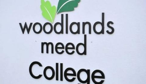 A black sign on a white school building reading Woodlands Meed College with a green leaf emblem above