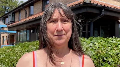 A woman with brown hair looking directly into the camera, with a serious expression. She is wearing a bright orange vest with straps and two golden necklaces.