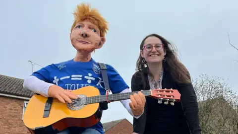 Sharon Kulesa Uma mulher - Sharon Kulesa - sorrindo para a câmera enquanto está ao lado de uma efígie de Ed Sheeran que está vestindo um top de Ipswich Town e segurando uma guitarra.
