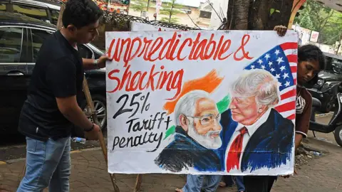 Ashish Vaishnav/Sopa Images/LightRocket via Getty Images two young people carry a handmade Prime Minister Narendra Modi and President Donald Trump with writing "Unpredictable and shocking 25% tariff + fine"