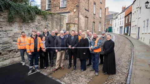 Durham County Council Group of people holding ribbon while standing on a cobbled street. They are all smiling. 