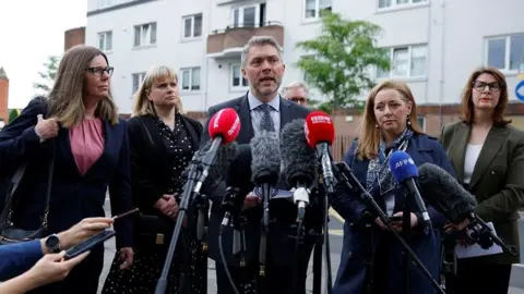 PA Media BBC NI director Adam Smyth (centre) in a grey suit, stood beside a blonde woman in black, a brown haired woman in navy and a man with grey hair and a navy suit, stood in front of numerous microphones