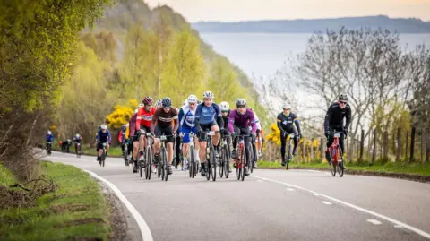 Paul Campbell A group of cyclists pedal up a hill, some of them off their saddles and standing as they push the pedals on their bikes. The road is flanked by trees and there is green, leafy woodland behind the group.