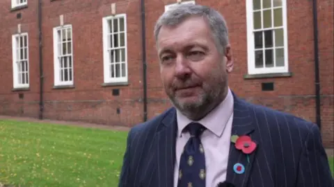 BBC A man with short grey hair and a black blazer with a navy tie and a remembrance poppy is stood near grass outside a brick building. He is looking away from the camera to the side.