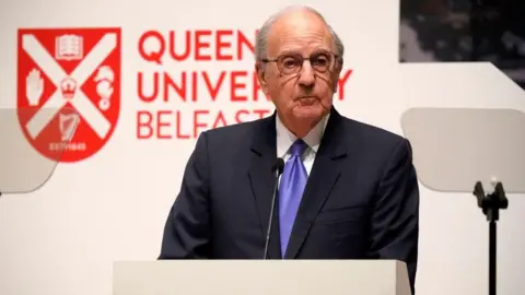 Getty Images George Mitchell, with short grey hair, wearing glasses, a navy suit, white shirt and purple tie. He is standing behind a white podium. There is a white wall behind him with Queen's University Belfast and the university logo in red. 