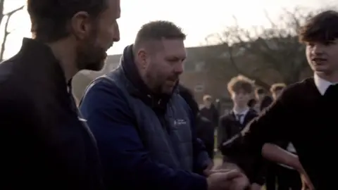 Gaze is speaking in a group of young people in school uniform while Gareth Southgate looks on