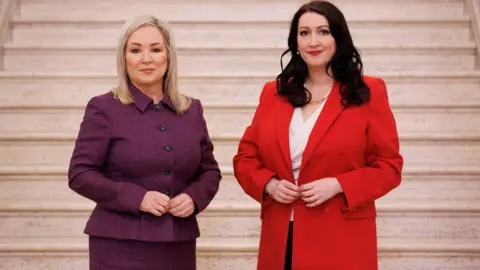 PA Media Michelle O'Neill wearing a purple suit, she has blonde hair. Emma Little-Pengelly in a red blazer and white top, she has black hair.