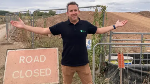 A man wearing a black polo shirt and brown trousers is standing in front of a large clay bund - there is also a red sign saying 'road closed'.
