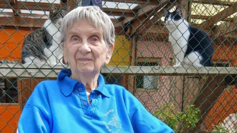 A woman wearing a blue sweatshirt stands in front of a wire‑mesh enclosure, with two cats sitting on a wooden platform behind the fence.
