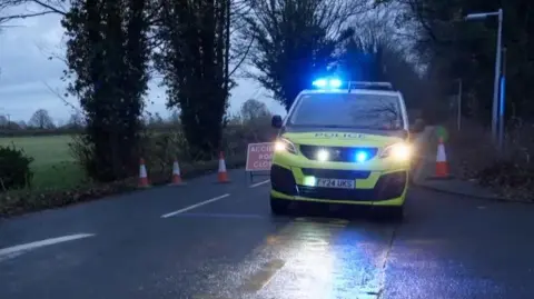 BBC A GV of a police car on a rainy country road in front of cones and a do not pass sign 