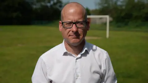 A bald man with rectangular black-rimmed glasses is looking directly into the camera. He is wearing a white shirt and the background is blurred but he is stood on a field with a goalpost in the background.