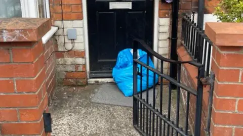 Krystal Selmi A blue parcel in front of a black front door. In the foreground is a red brick wall and a black iron gate.