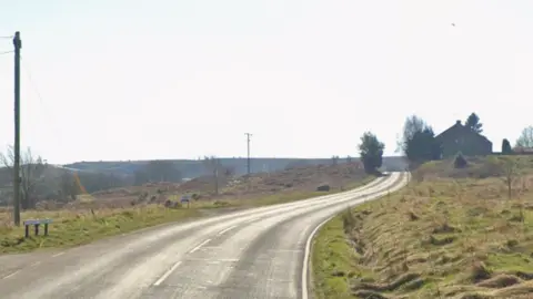 A quiet bending country road with long grass on either side. In the distance is a house