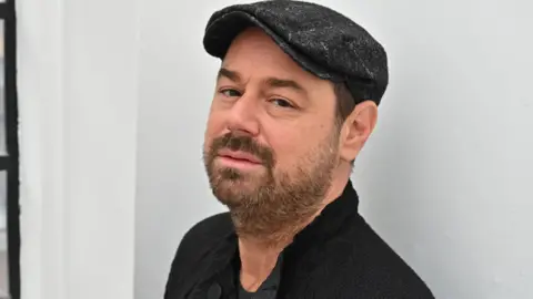 Headshot Brit True dari Danny Dyer memandangi kamera dengan topi hitam, Anda dapat melihat bagian atas kardigan hitam dan kemeja hitam. Dia memiliki ekspresi hangat di matanya, mulutnya netral. Ada dinding putih di belakangnya. 