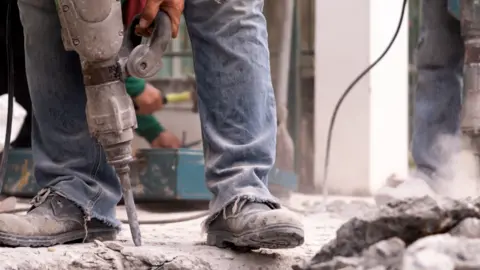 A builder uses equipment to drill into concrete. He is wearing jeans and old boots.