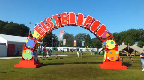 Sign with Eisteddfod spelt out in red over an arch with coloured green, blue, yellow and orange circles festooning the arch. In the background you can see festival flags in blue and yellow and orange and people walking around festival tents