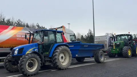 Two tractors, one blue and one green, one in front of the other on a road in Ireland with a fuel tanker beside one of them. The blue tractor is towing a blue trailer.