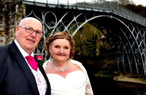 Cancer Research UK A groom and bride are dressed up near the Iron Bridge in Shropshire. They are on the left of the photo with the man wearing glasses and the woman is in a bridal dress