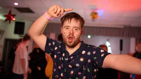 BBC Man is wearing a Christmas themed polo shirt. He is wearing a pink glow stick bracelet and has his hand in the air, finger pointing while he's dancing. He has glitter on his cheeks. He has brown, spiky hair. 