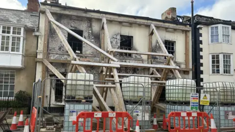 Burned building with scaffolding and large wooden beams propping it up. The front of the building is largely bare after the fire, and there are orange safety barriers and cones at street level