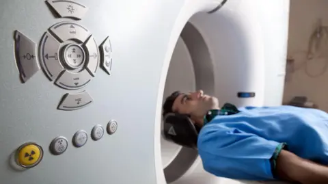 A male patient, wearing a blue hospital gown, lies on a CT scanner. The dials of the scan machine are shown on the left of the picture.