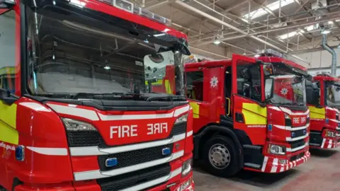 Three Cheshire Fire and Rescue Service branded fire engines parked alongside one another, in a fire station. The middle fire engine has its door open.