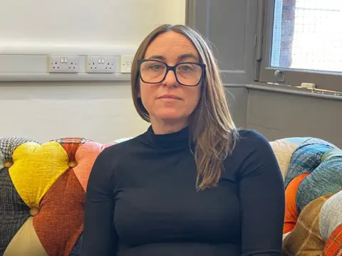 BBC News A woman sits on a sofa with a brightly coloured diamond pattern. She has brown hair and wears black glasses and a black high neck jumper 