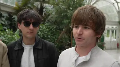 Dan Parker, wearing a white top, and Oisin McAvoy, wearing sunglasses and a black jacket over a white top, are being interviewed in Sefton Park Palm House. They are surrounded by trees and shrubs in a spacious white atrium