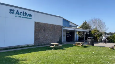 Les Quennevais Sports Centre. A modern single-story building with a front wall comprising of a mixture of concrete and stone, with an entrance featuring an solid awning above it on the right-hand side. In front is a lawn with two picnic benches   