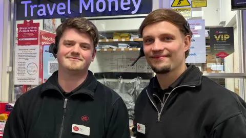 Two young men in Post Office uniforms - black fleeces and shirts - with white name badges. Behind them are bags of post and lots of notices with the Post Office's red branding.
