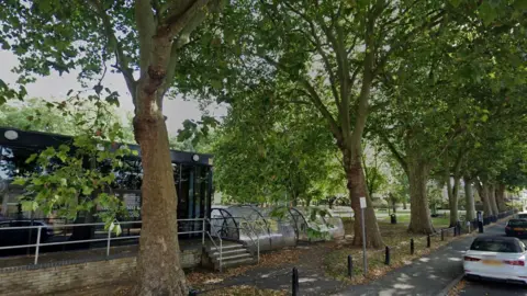 A row of tall trees are in front of a single storey glass fronted building. A covered bike rack is next to it. In front of the trees is a short metal fence, a path and a road with parked cars. 